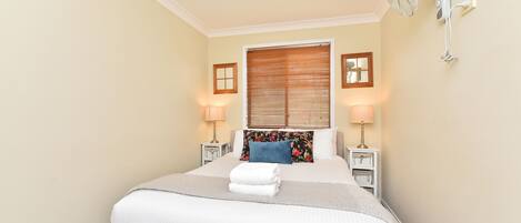 A hotel room with a bed, two bedside lamps, a window with blinds, and a wall-mounted fan.