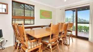 A dining area with a wooden table and chairs, large windows with blinds, a green wall art piece, and a view of greenery outside. - Silver Springs Cottage Hunter Valley Retreat (Rothbury)
