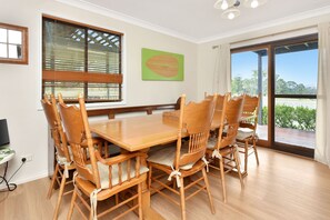 A dining area with a wooden table and chairs, large windows with blinds, a green wall art piece, and a view of greenery outside.