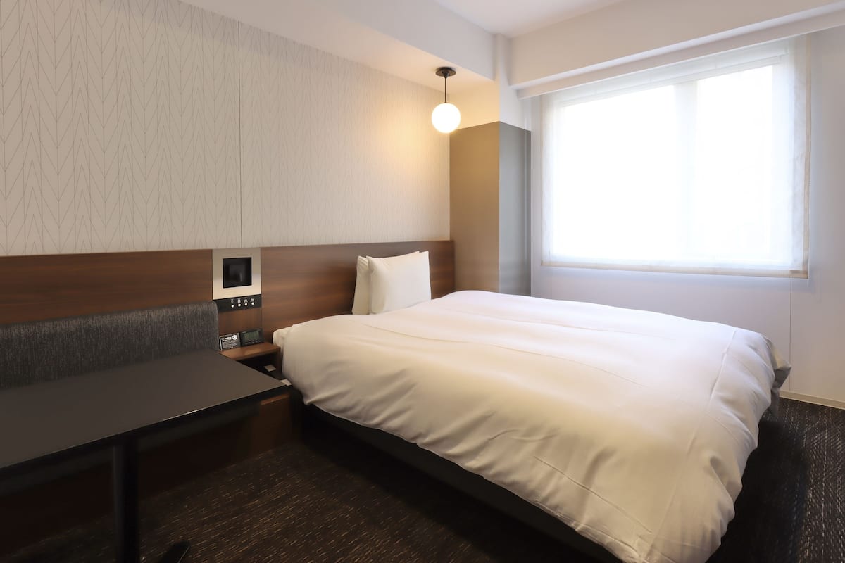 Une chambre d’hôtel équipée d’un lit, d’un bureau, d’une table de chevet, d’un panneau de commande fixé au mur et d’une fenêtre avec des stores.