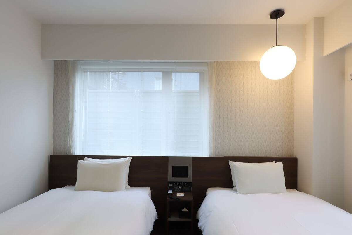 Une chambre d’hôtel avec deux lits, une table de chevet, un panneau de commande et une grande fenêtre avec des stores.