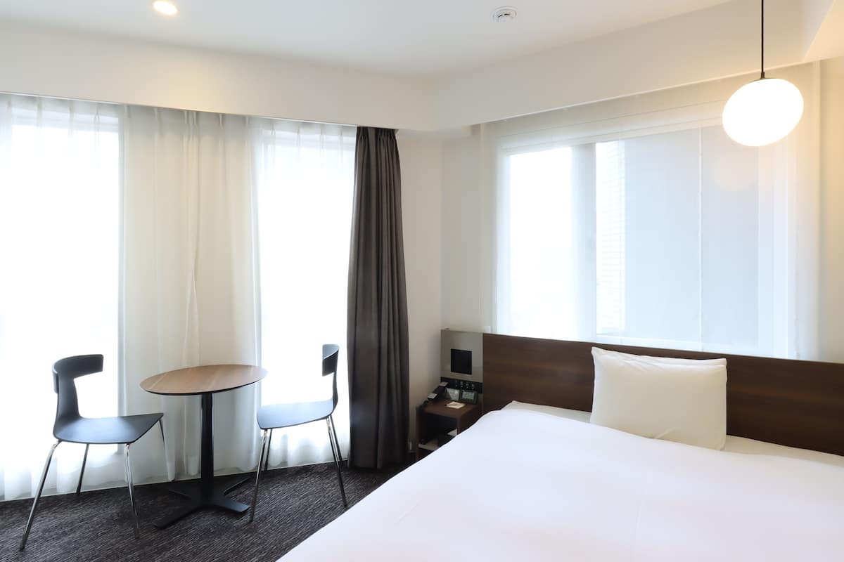 Une chambre d’hôtel avec un grand lit, une petite table, deux chaises, une table de chevet et une fenêtre avec des rideaux.