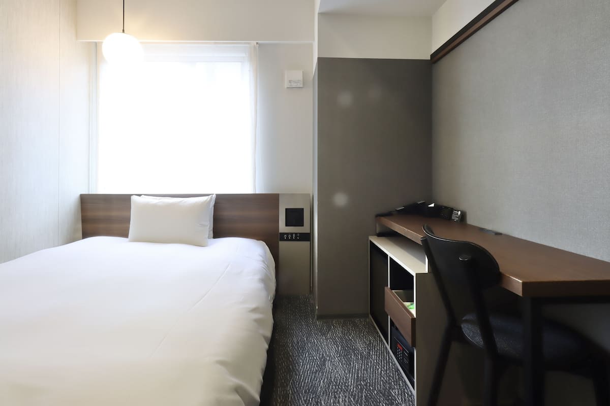 Une chambre d’hôtel équipée d’un lit, d’un bureau, d’une chaise, d’une lampe de bureau et d’une fenêtre avec des rideaux.