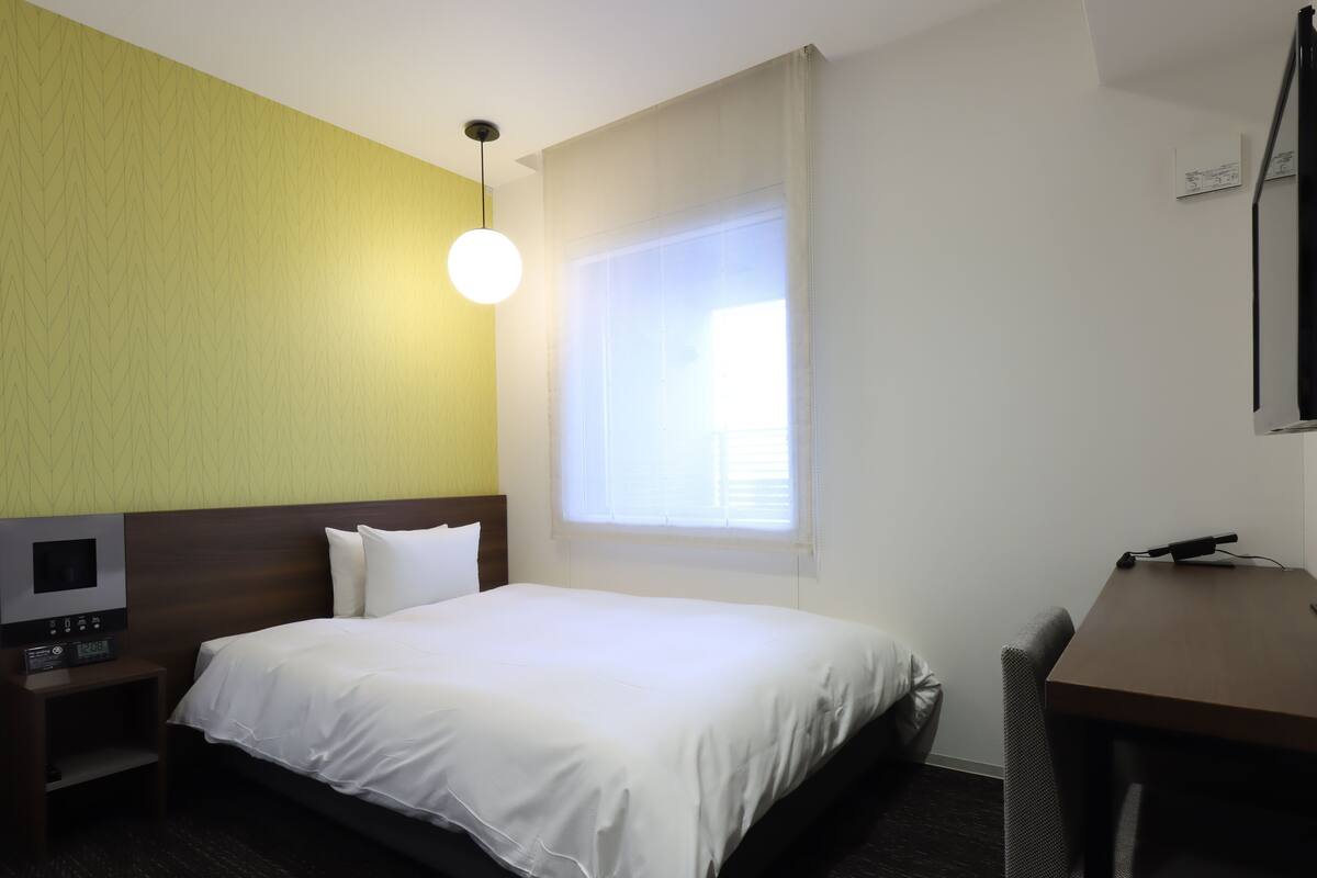 Une chambre d’hôtel équipée d’un lit, d’un bureau, d’une chaise, d’une télévision et d’une fenêtre avec des stores.