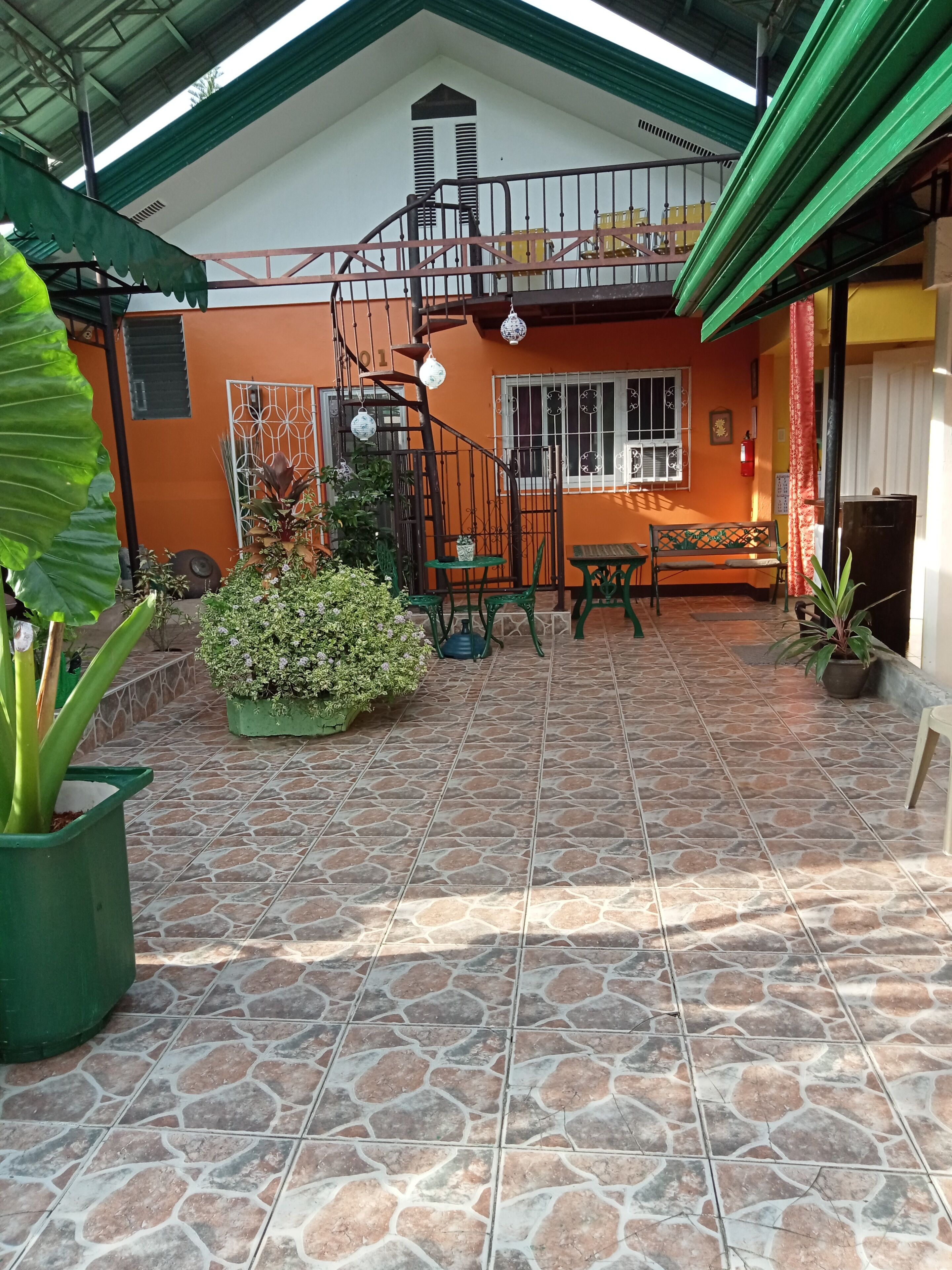 A covered patio area with a tiled floor, a green bench, a small table, and potted plants.