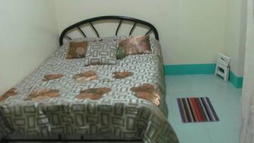 A single bed with a patterned bedspread and pillows, a black metal headboard, a small white radiator, and a striped rug on a light-colored floor.