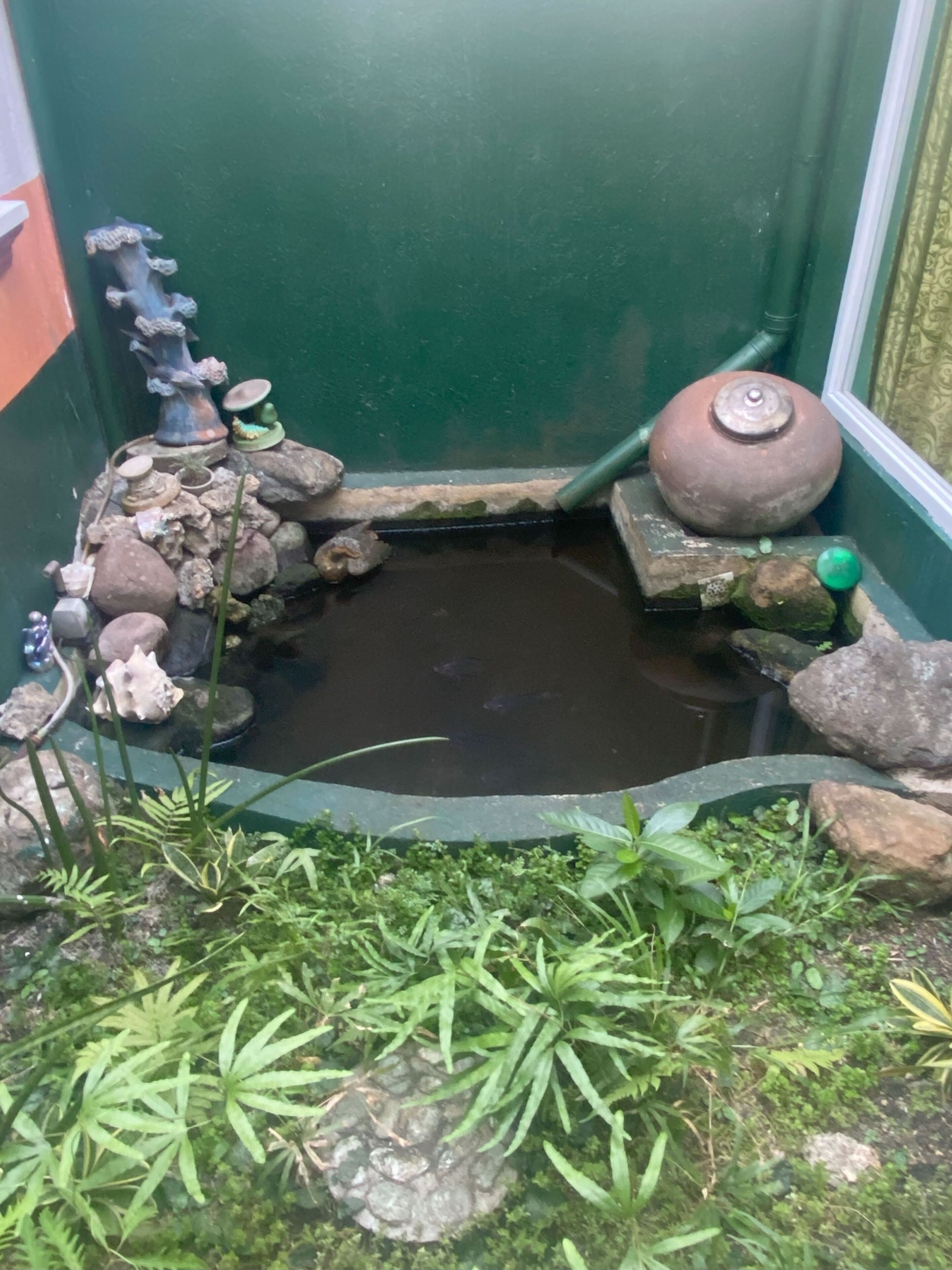Un pequeño estanque con rocas, plantas, una bomba de agua y un adorno sobre un pedestal.