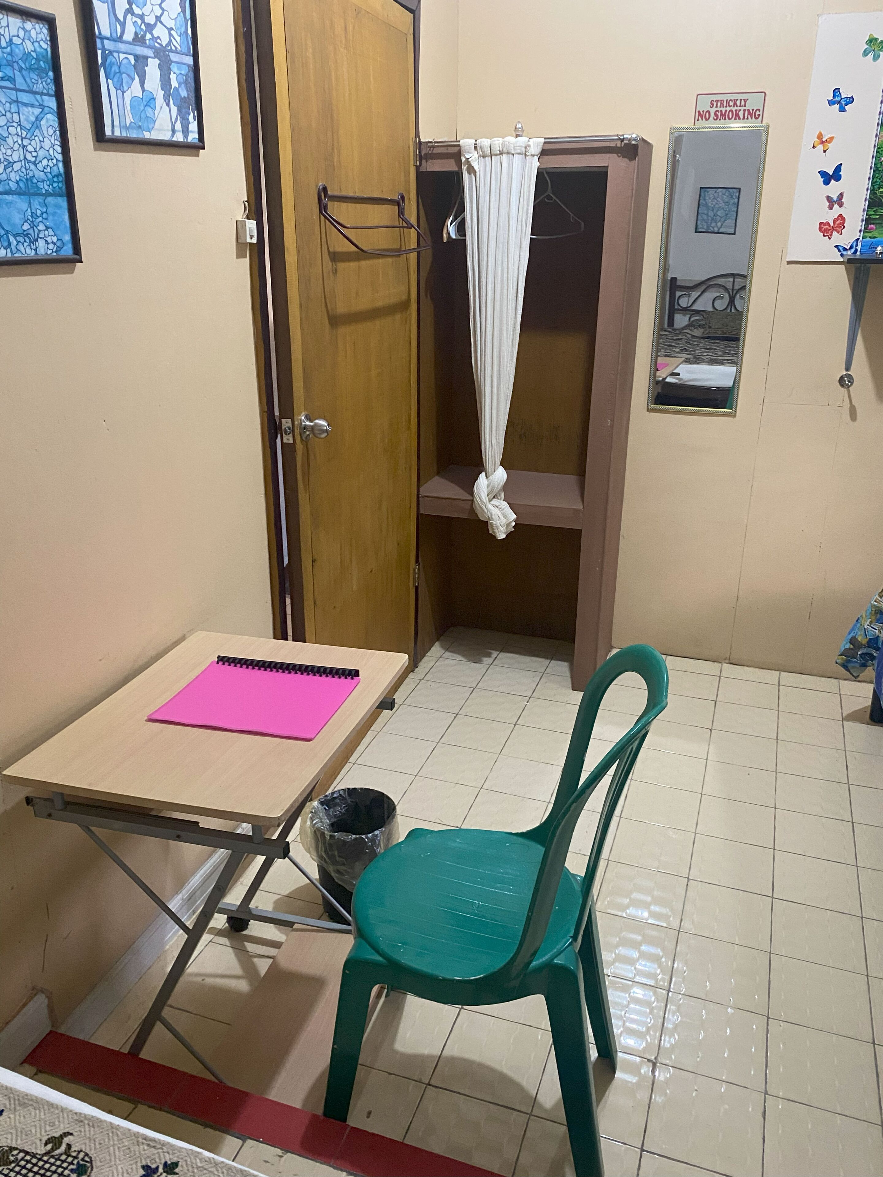 A small room with a wooden door, a green plastic chair, a table with a pink notebook, a black trash bin, and a white curtain.