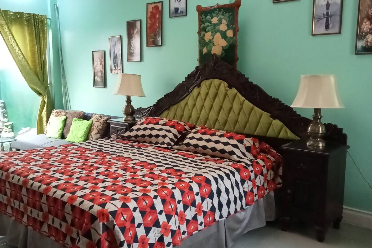 A bedroom with a bed, two bedside tables, a nightstand, a window with curtains, and wall-mounted artwork.