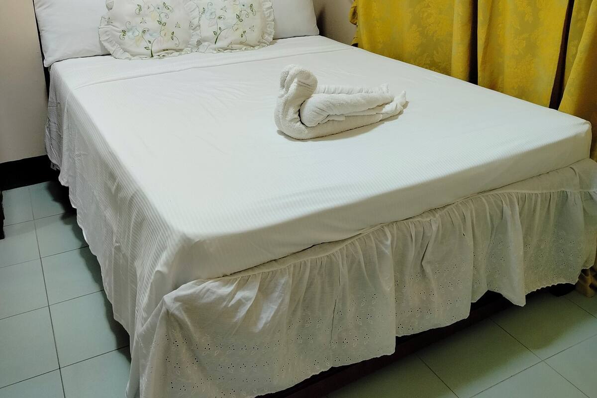 A neatly made bed with a white bedspread and a decorative pillow, a yellow curtain partially drawn, and a painting of a beach scene on the wall.
