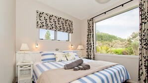 A bedroom with a large bed, two bedside lamps, a window with curtains, and a view of greenery outside.