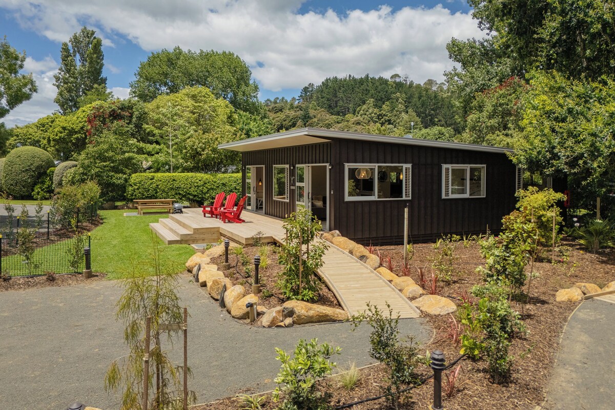 A modern cabin with a covered patio, surrounded by a well-maintained garden and a forested backdrop.