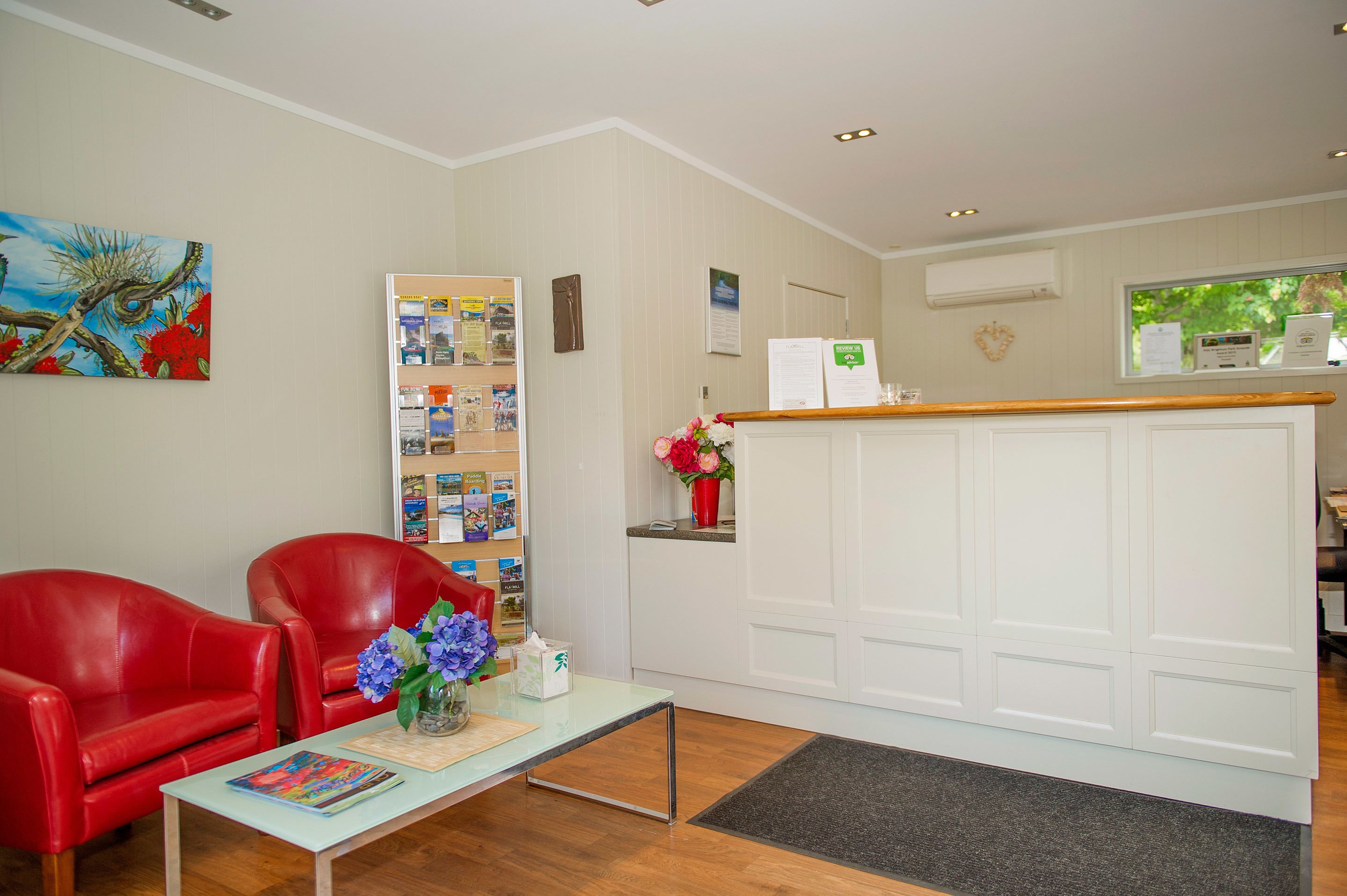 A reception area with a white reception desk, red leather chairs, a small table with a vase of flowers, and a bulletin board with various notices.
