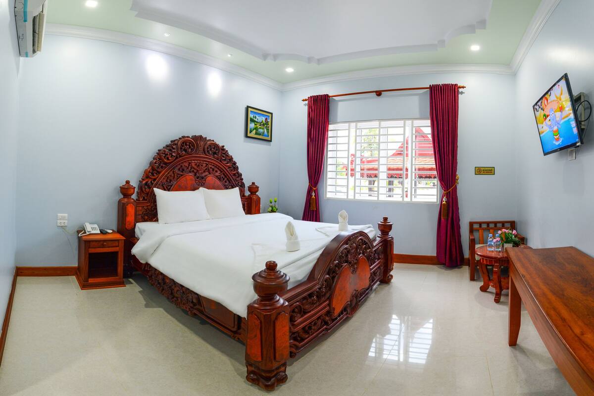 A bedroom with a large wooden bed, a bedside table, a nightstand, a TV mounted on the wall, and a window with curtains.