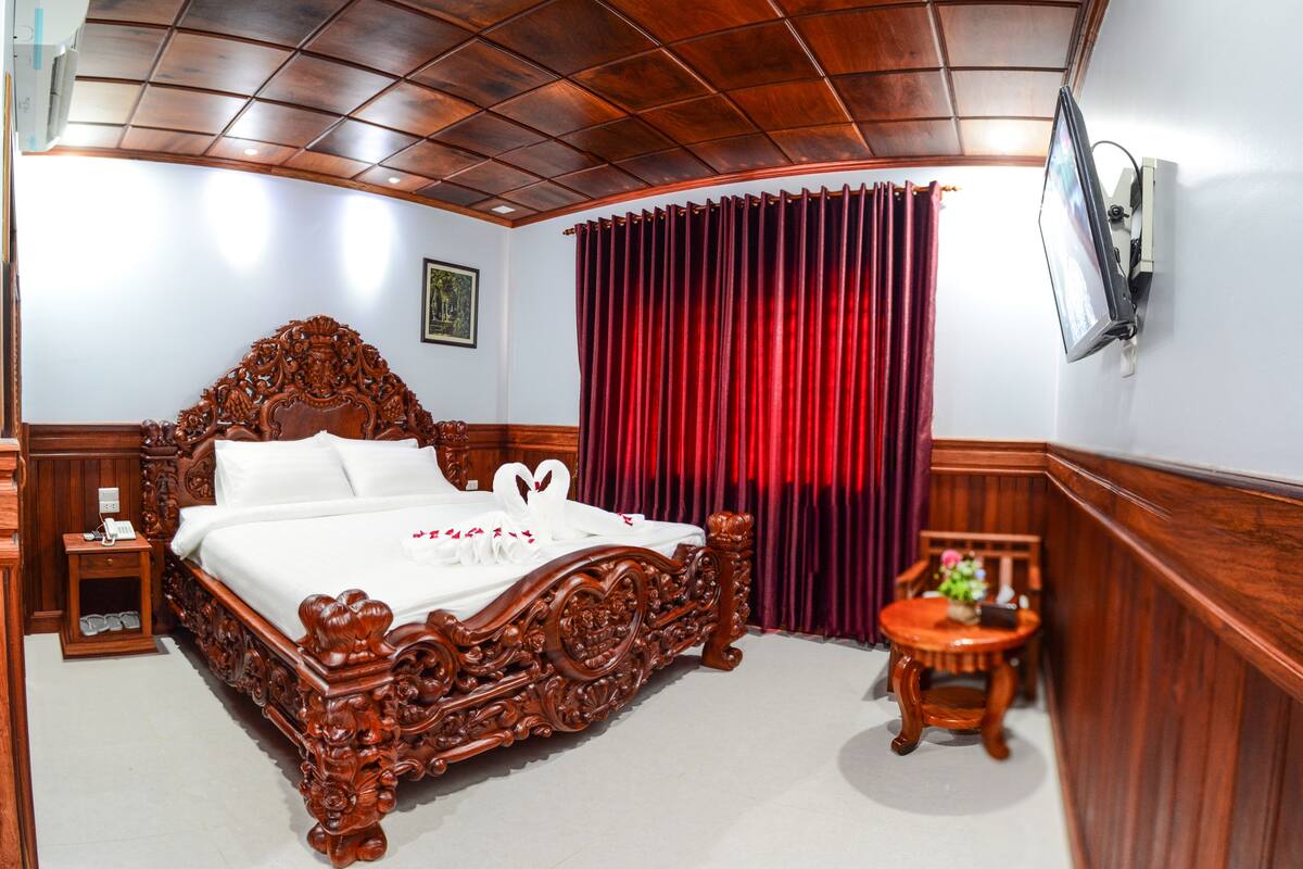A bedroom with a wooden bed, a red curtain, a TV mounted on the wall, and a small wooden table with a plant.