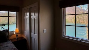 A bedroom with a bed, a nightstand with a lamp, a window with a view of a lake, and a door.