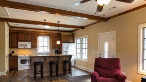 A spacious kitchen with wooden cabinets, a central island with stools, a ceiling fan, and a red armchair in a living area with hardwood floors.