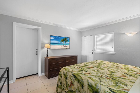 A bedroom with a bed, a dresser, a painting of a beach scene, and a window with blinds.