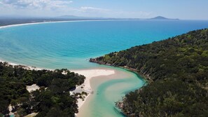 An aerial view of a coastal area with a sandy beach, clear turquoise waters, and dense green forest. - Hat Head Holiday Park (Hat Head)