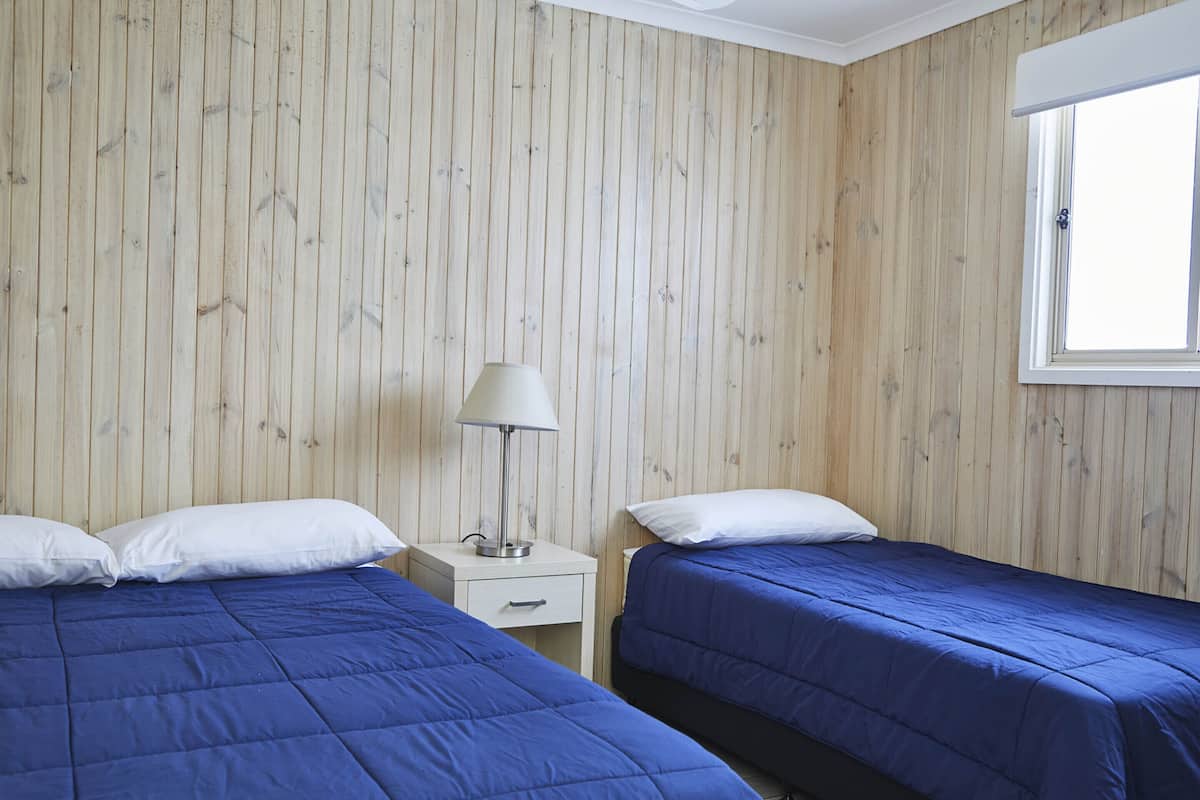 A room with two beds, wooden paneling on the walls, a nightstand with a lamp, and a window.