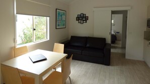 A room with a dining table, chairs, a black leather sofa, a framed picture on the wall, and a window with blinds.