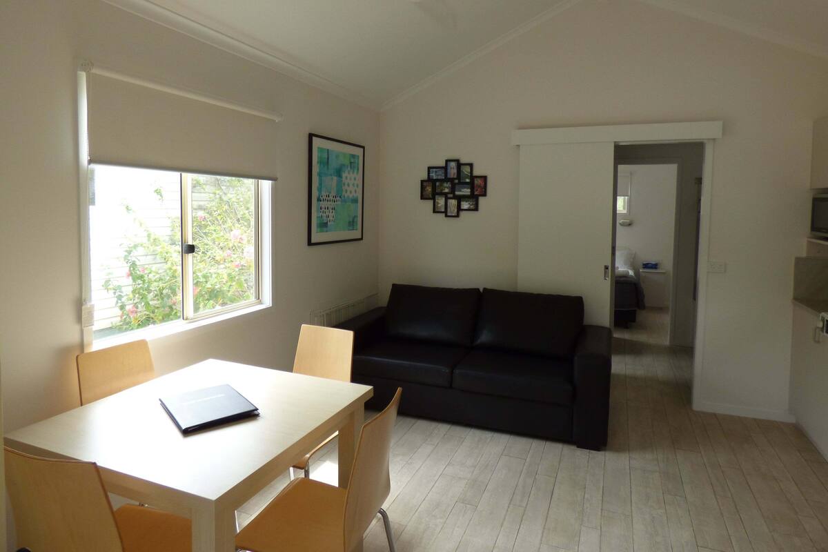 A room with a dining table, chairs, a black leather sofa, a framed picture on the wall, and a window with blinds.