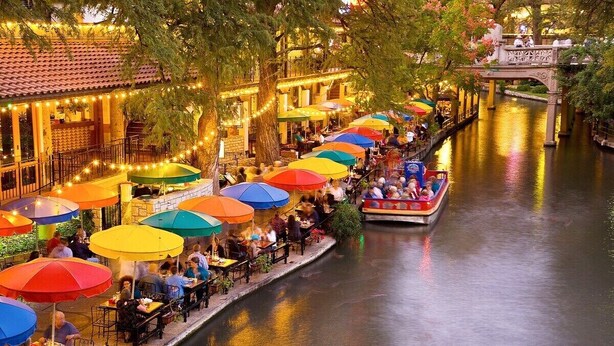 Een eetgedeelte aan de rivier met buiten zitplaatsen en parasols, een boot en een brug op de achtergrond.