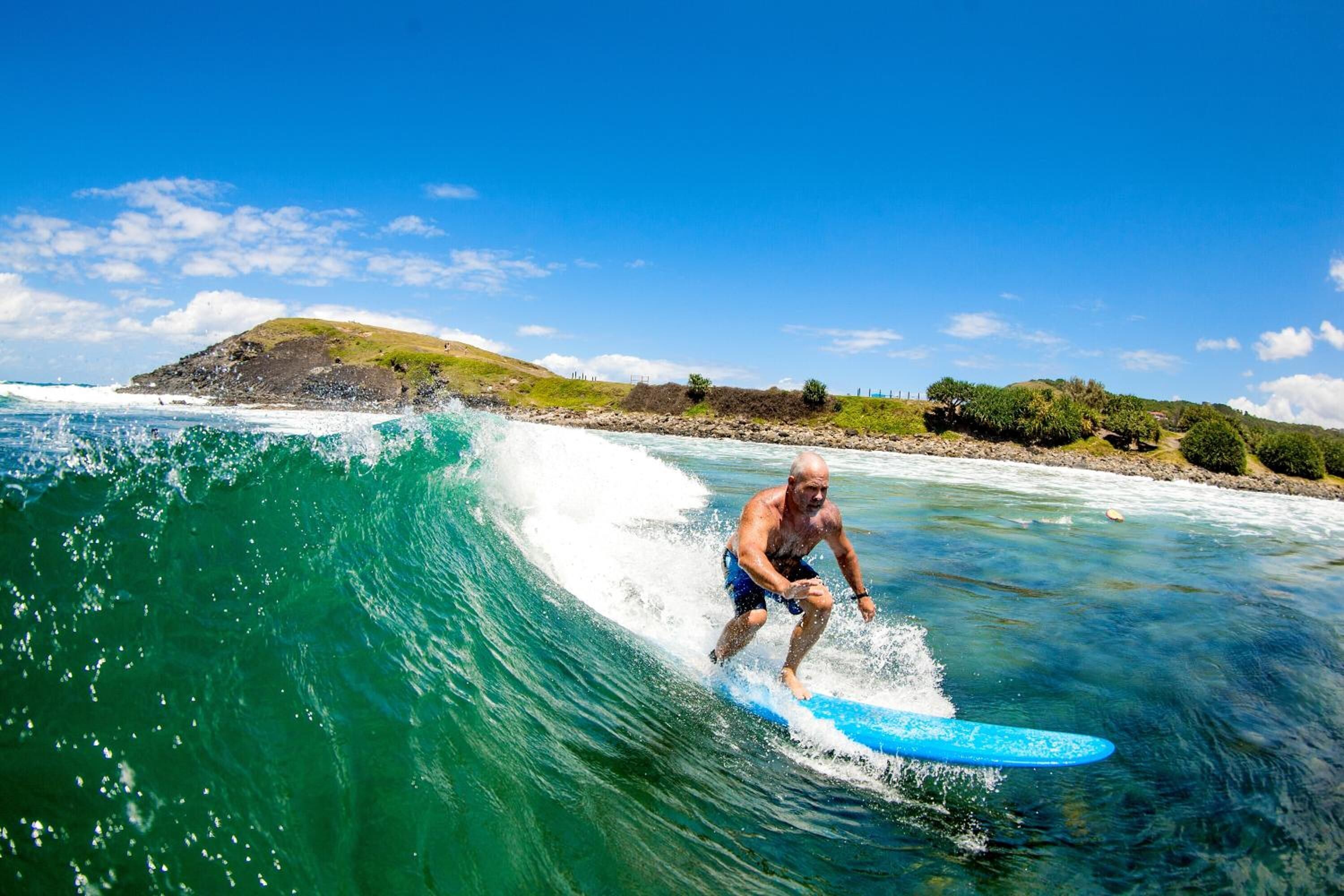 A man surfing on a clear day with a view of a beach and a hill in the background.