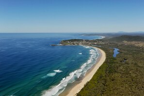 An aerial view of a coastal area with a beach, clear blue waters, and a small settlement with buildings and greenery. - Crescent Head Holiday Park (Crescent Head)