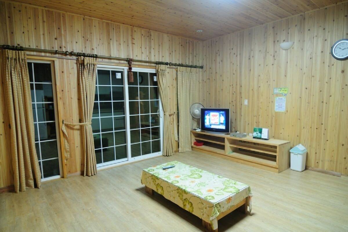 Une pièce aux murs en bois, une porte-fenêtre coulissante, un lit recouvert d’une courtepointe à motifs floraux, un téléviseur posé sur un meuble en bois et une horloge au mur.