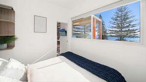A bedroom with a bed, a window with a view of trees, a shelf with a plant, and a door leading to another room. - Horseshoe Bay Holiday Park (South West Rocks)