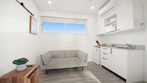 A modern, minimalist room with a sofa, a wooden bench, a kitchen area with white cabinets, and a window providing natural light. - Horseshoe Bay Holiday Park (South West Rocks)
