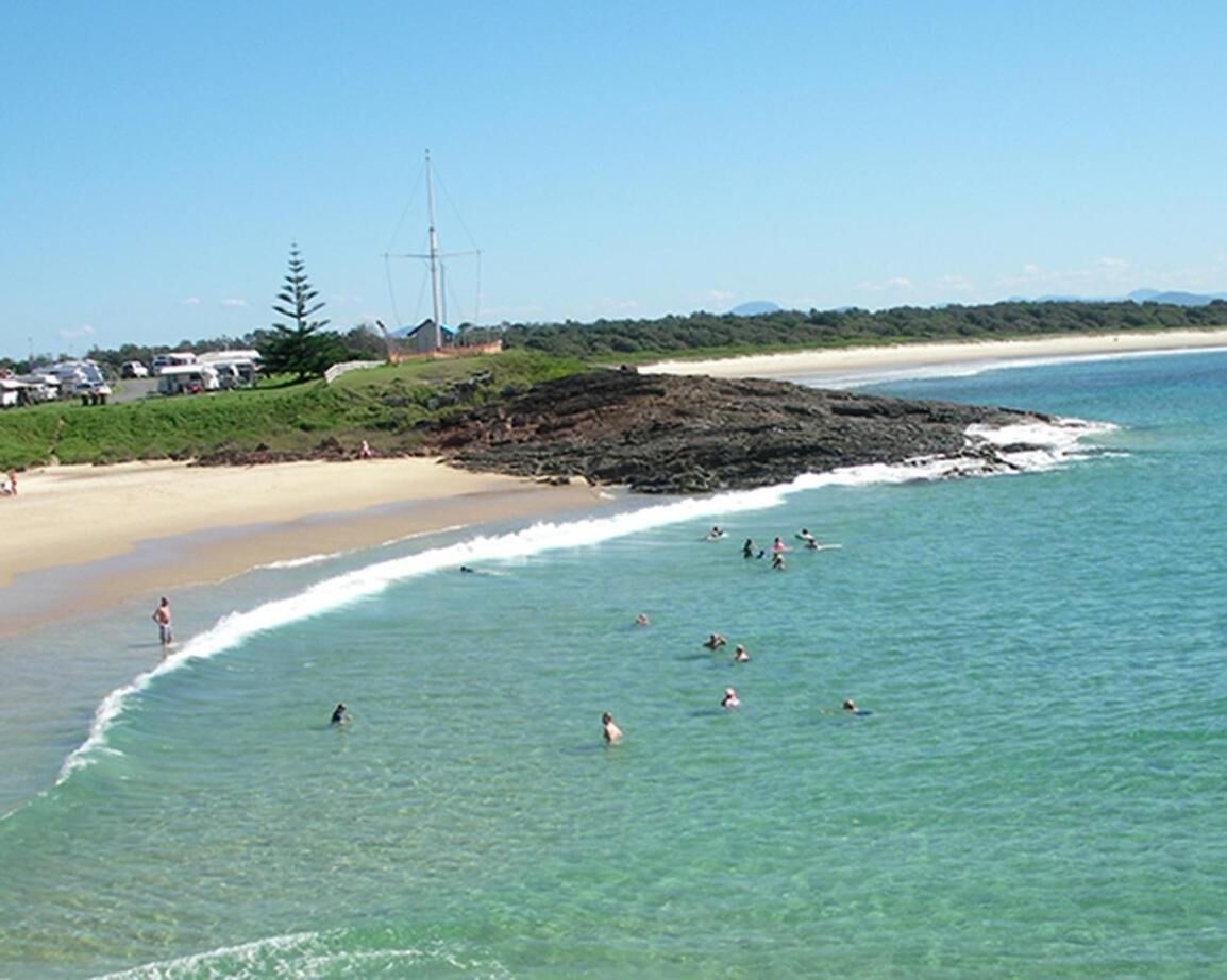 A beach with clear turquoise water, people swimming and sunbathing, and a sandy shore with a few buildings in the background.