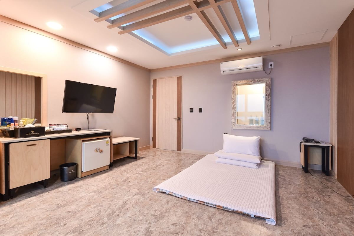 A hotel room with a bed, a desk with a flat-screen TV, a minibar, and an air conditioning unit.