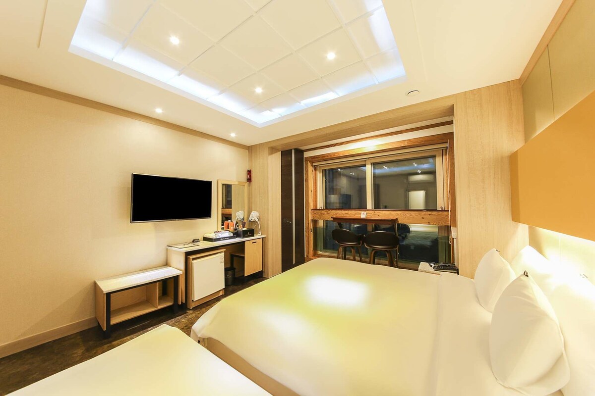 A modern hotel room with a large bed, a desk with a flat-screen TV, a vanity with a mirror, and a large window.