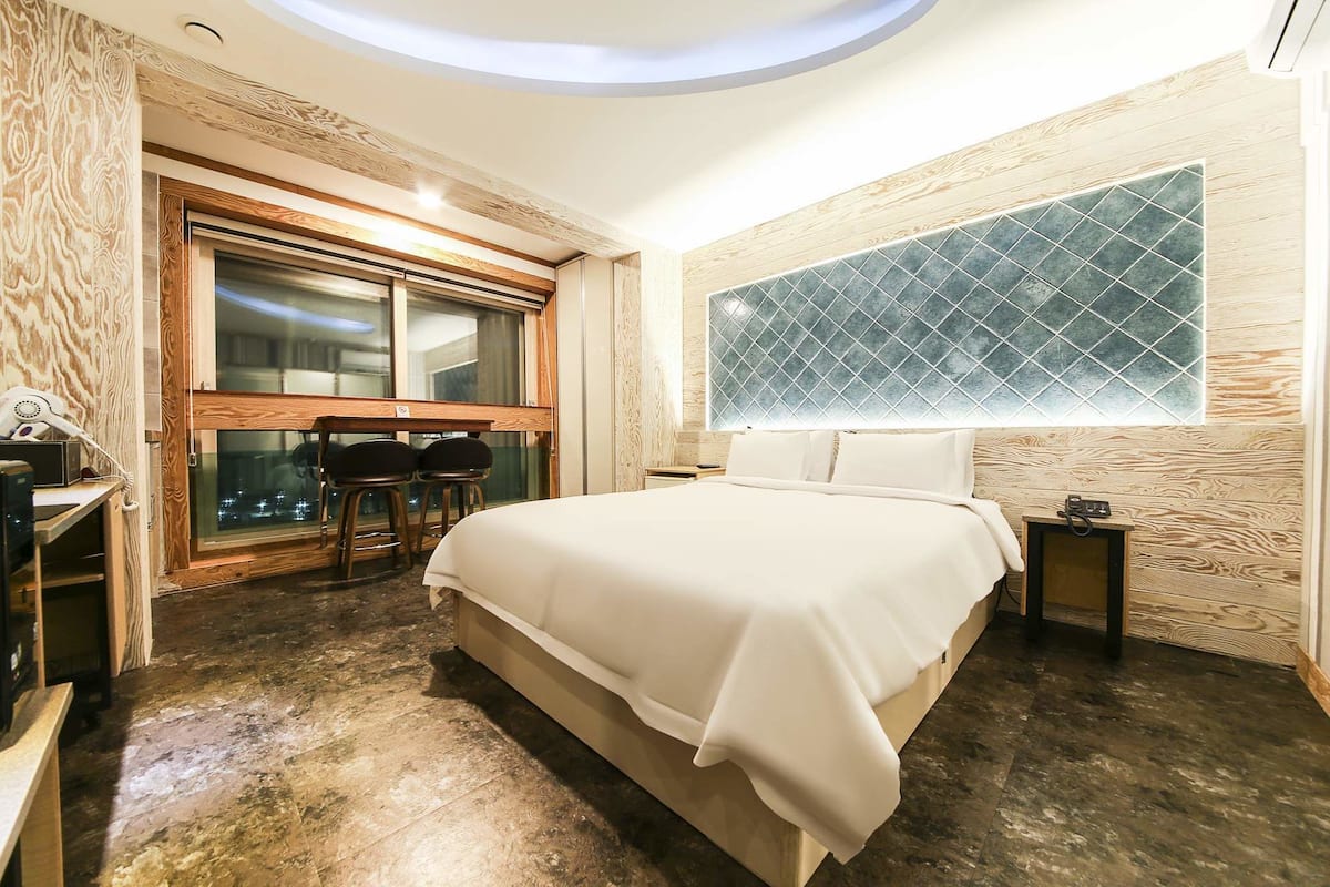 A modern hotel room with a large bed, a small desk, a chair, and a window with a geometric pattern.