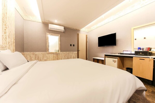 A hotel room with a bed, a desk with a flat-screen TV, a mirror, and a small refrigerator.