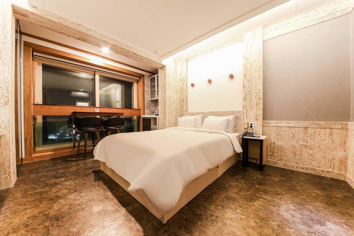 A hotel room with a large bed, a desk with two chairs, a window with a view of the city, and a nightstand with a lamp.
