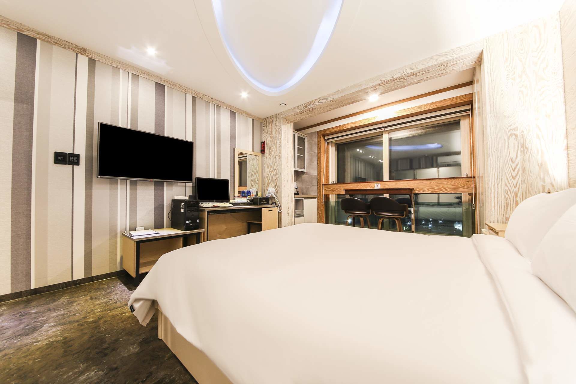 A hotel room with a large bed, a desk with a computer, a flat-screen TV, and a window with a view of the city at night.