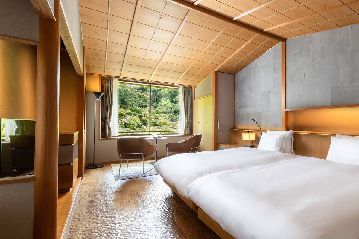 A hotel room with two beds, a desk, chairs, a TV, and a large window with a view of trees.