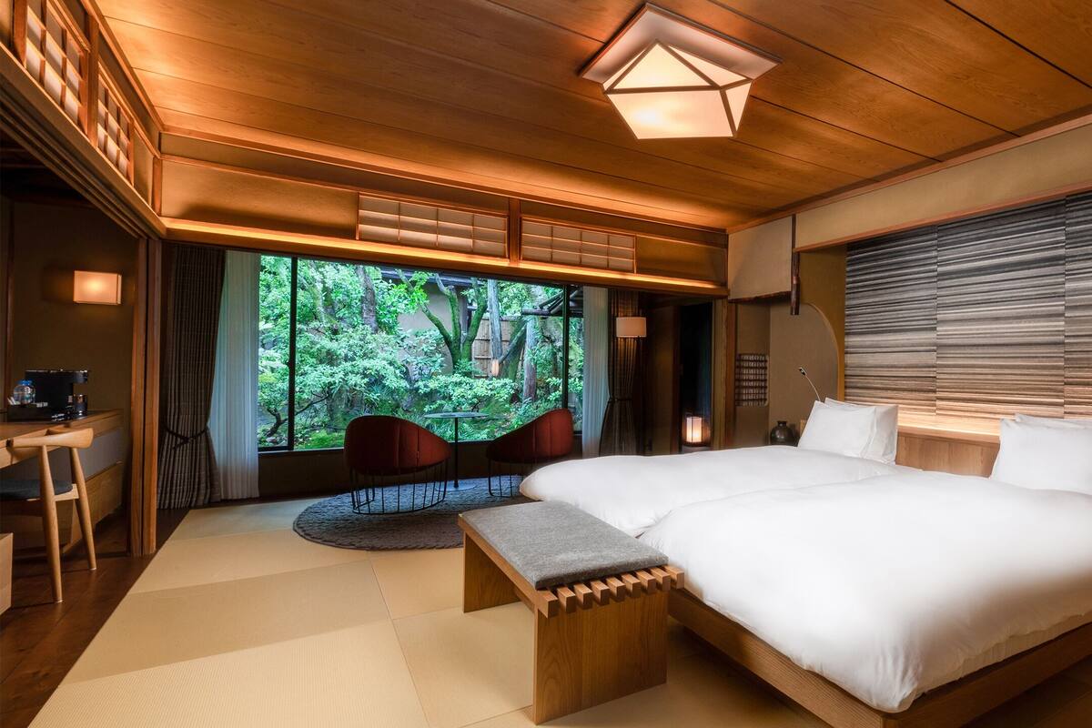 A modern bedroom with a large bed, wooden accents, a view of greenery, and a desk with a chair.