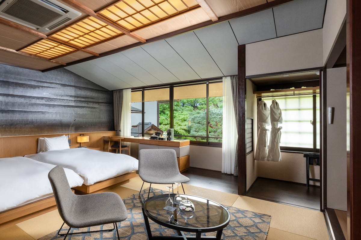 A hotel room with two beds, a sitting area with a glass table, and a view of greenery outside.