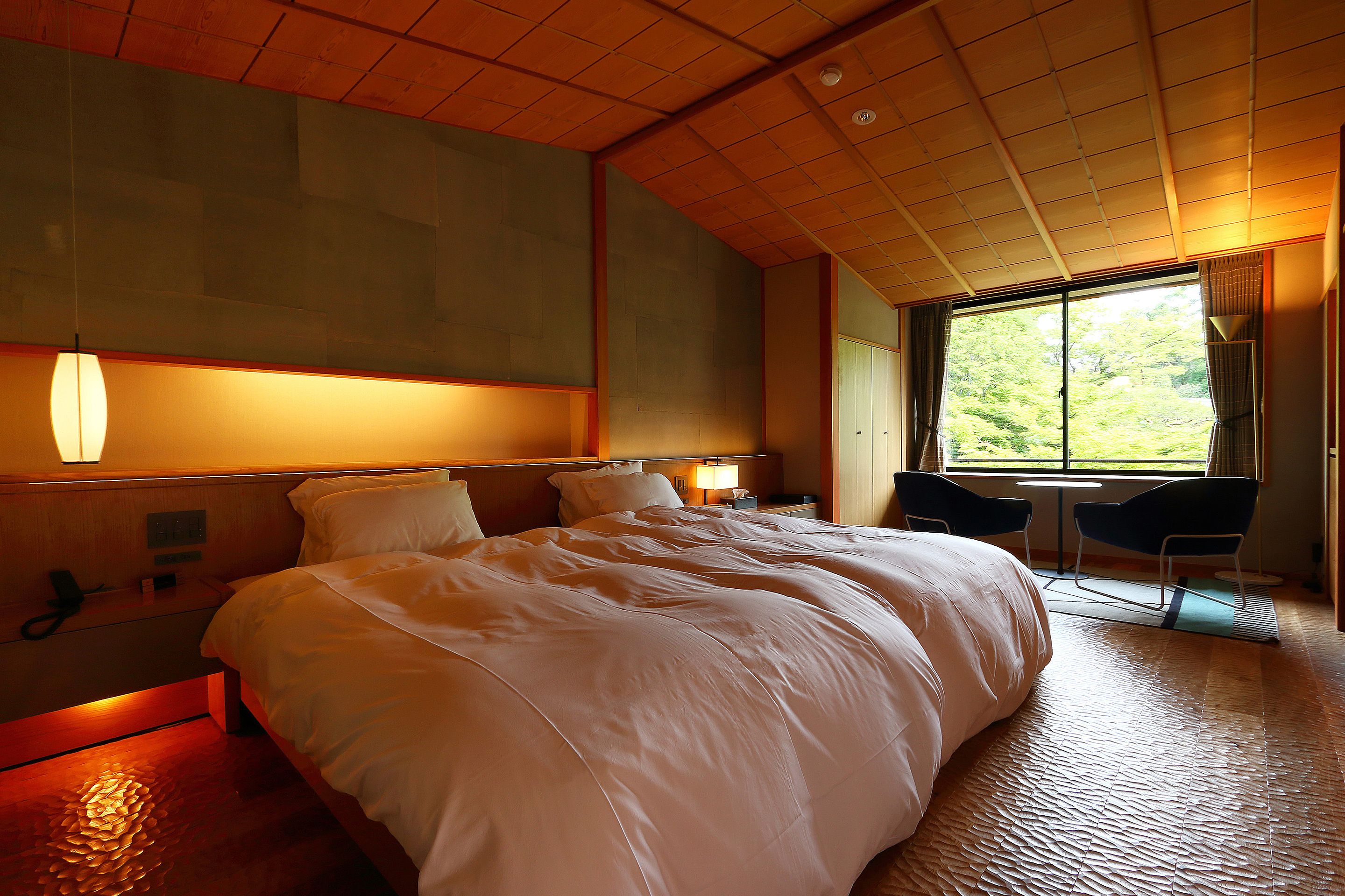 A large bed with a wooden headboard, a bedside table with a lamp, and a window with a view of greenery.
