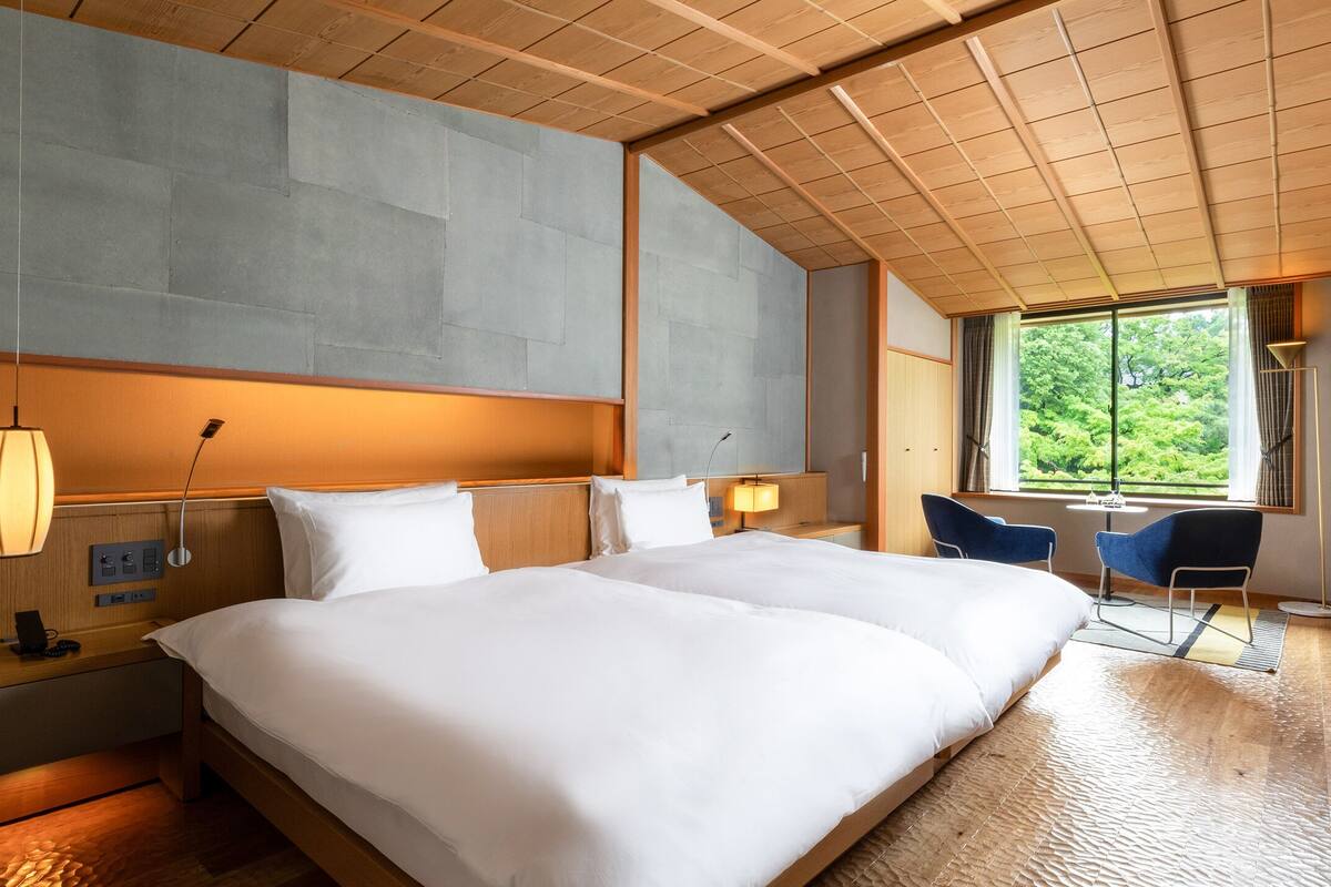 A hotel room with a large bed, bedside tables, a desk, a chair, and a window with a view of greenery.