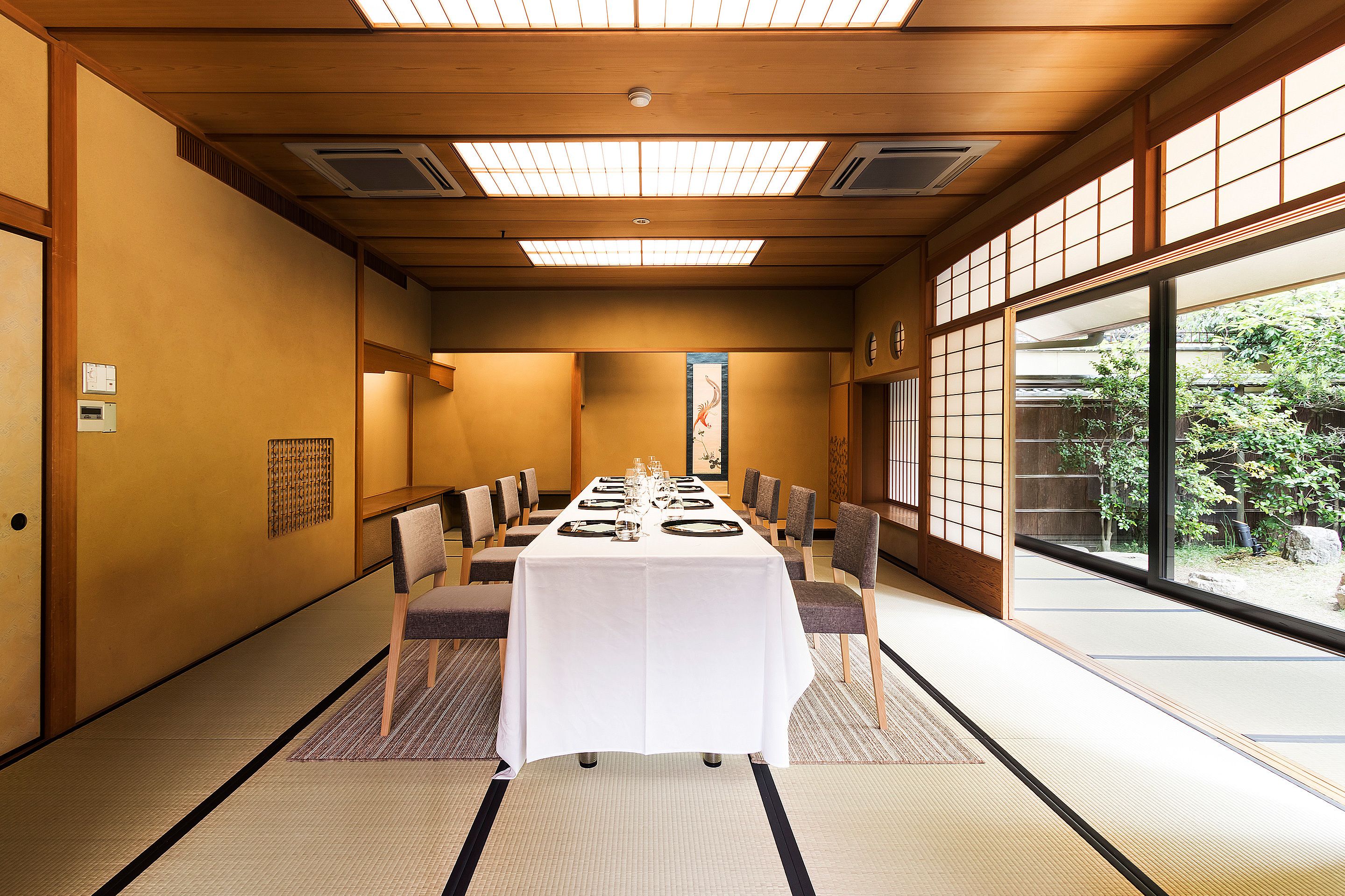 A traditional Japanese-style room with tatami flooring, a low table set for dining, and large windows offering a view of a garden.