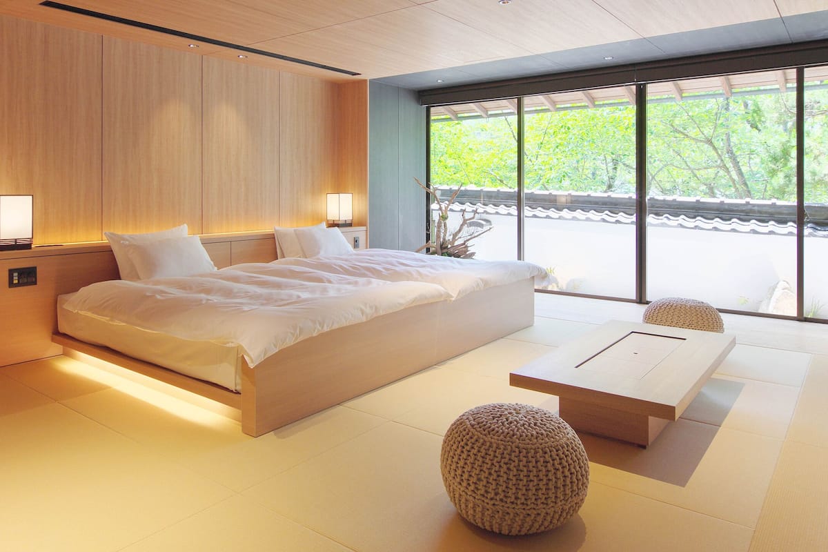 A modern bedroom with a large bed, bedside lamps, a wooden headboard, tatami flooring, and a view of greenery through large windows.