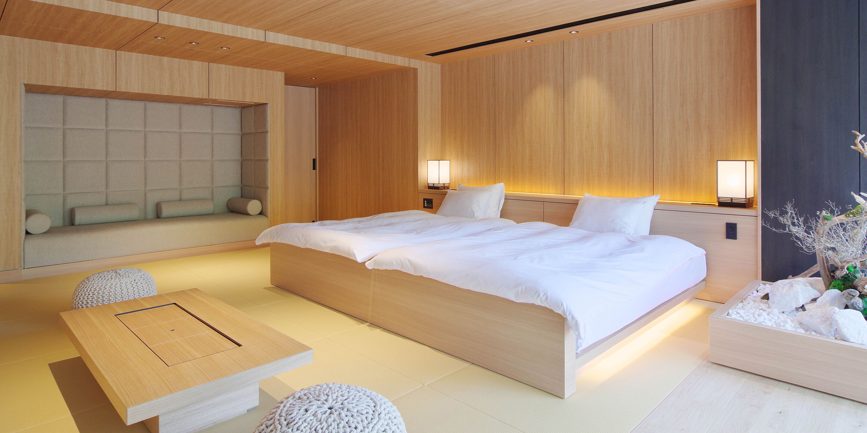 A modern bedroom with a large bed, bedside lamps, a wooden headboard, and a small table with a cushion.