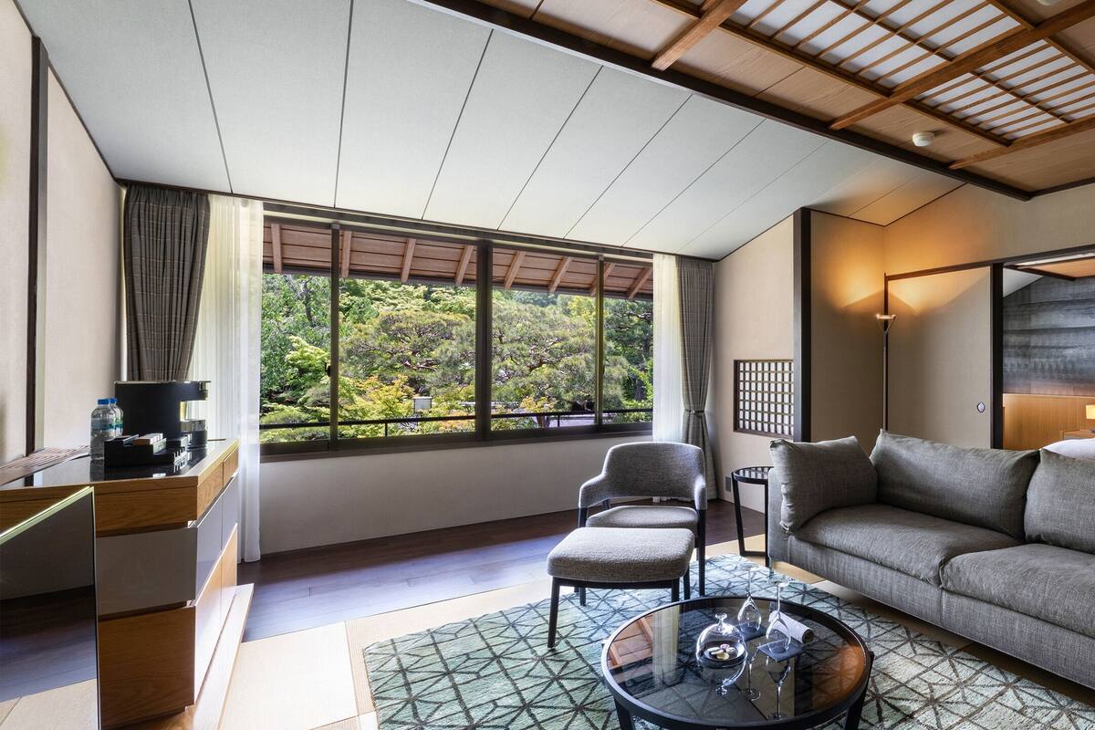 A modern living room with a sofa, armchair, and coffee table. There is a large window with a view of greenery outside.