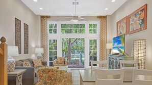 Un salon spacieux doté d’une grande fenêtre, d’un ventilateur de plafond et d’un coin repas avec des chaises et une table blanches.