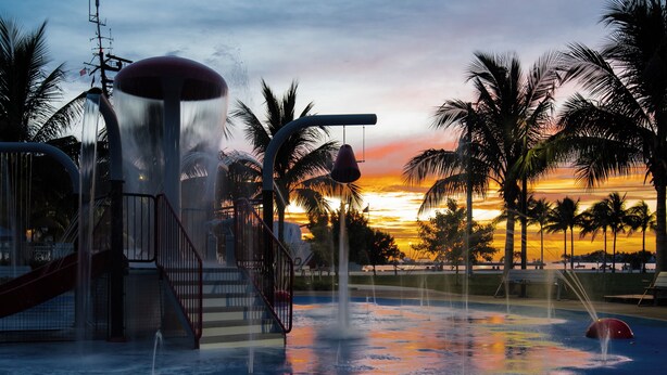 Un parc aquatique avec un toboggan, une fontaine et des palmiers, le tout baigné par les couleurs du coucher de soleil.